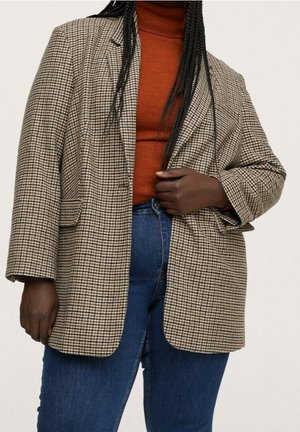 Personne portant un blazer à carreaux beige et marron sur un pull à col roulé couleur rouille et un jean bleu, tenant le blazer fermé.
