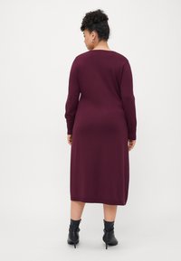 Woman with curly hair wearing a long-sleeve, knee-length burgundy dress and black ankle boots, standing facing away against a plain background.