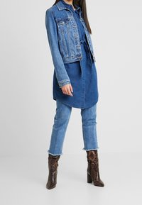 Giubbotto di jeans, blu medio, taglio classico con tasche sul petto sopra un vestito camicia blu, abbinato a jeans in denim chiaro e stivali con tacco in pelle di serpente marrone scuro.