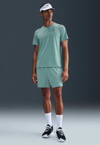Atletische man in een lichtgroen matching shorts en t-shirt, witte sokken, zwart-witte sneakers en een witte Nike visor, staande op een grijze vloer.
