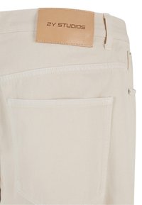 Beige denim pants with a smooth texture, featuring a brown leather tag that reads "2Y STUDIOS" and a subtle back pocket design.