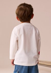 Biela dlhá rukávová košeľa s okrúhlym výstrihom, s červeným detailom na ľavom rukáve. Mäkka textúra, voľný strih, v páre s džínsovými šortkami.