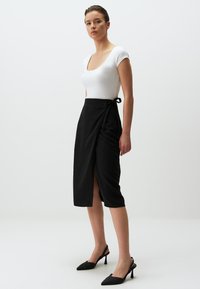 Zwarte wikkelrok met een strik in de taille en een split. Gecombineerd met een wit kortblauw, aansluitend bovenstuk en zwarte puntige hakken.