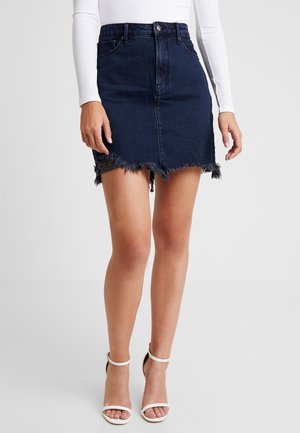 Femme portant une jupe en denim bleu foncé à ourlet effiloché, un haut blanc à manches longues, et des sandales blanches à talons hauts sur un fond uni.