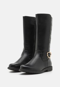 Bottes noires montantes en cuir lisse, avec un accent tressé et un détail en matériel en forme de cœur doré sur le côté.