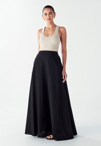 Ženska s temnimi lasmi, oblečena v bež brezrokavnik in dolgo črno krilo, stoji pred enotno belo ozadjem.