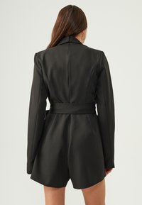 Black tailored blazer dress with long sleeves, structured shoulders, belted waist, and smooth fabric. Features minimalist design and clean lines.