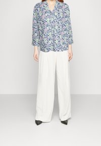 Leichtgewichtige Bluse mit Blumenmuster in Blau, Grün und Pink; kombiniert mit hochgeschnittenen, weit geschnittenen weißen Hosen; schwarze spitze Schuhe.