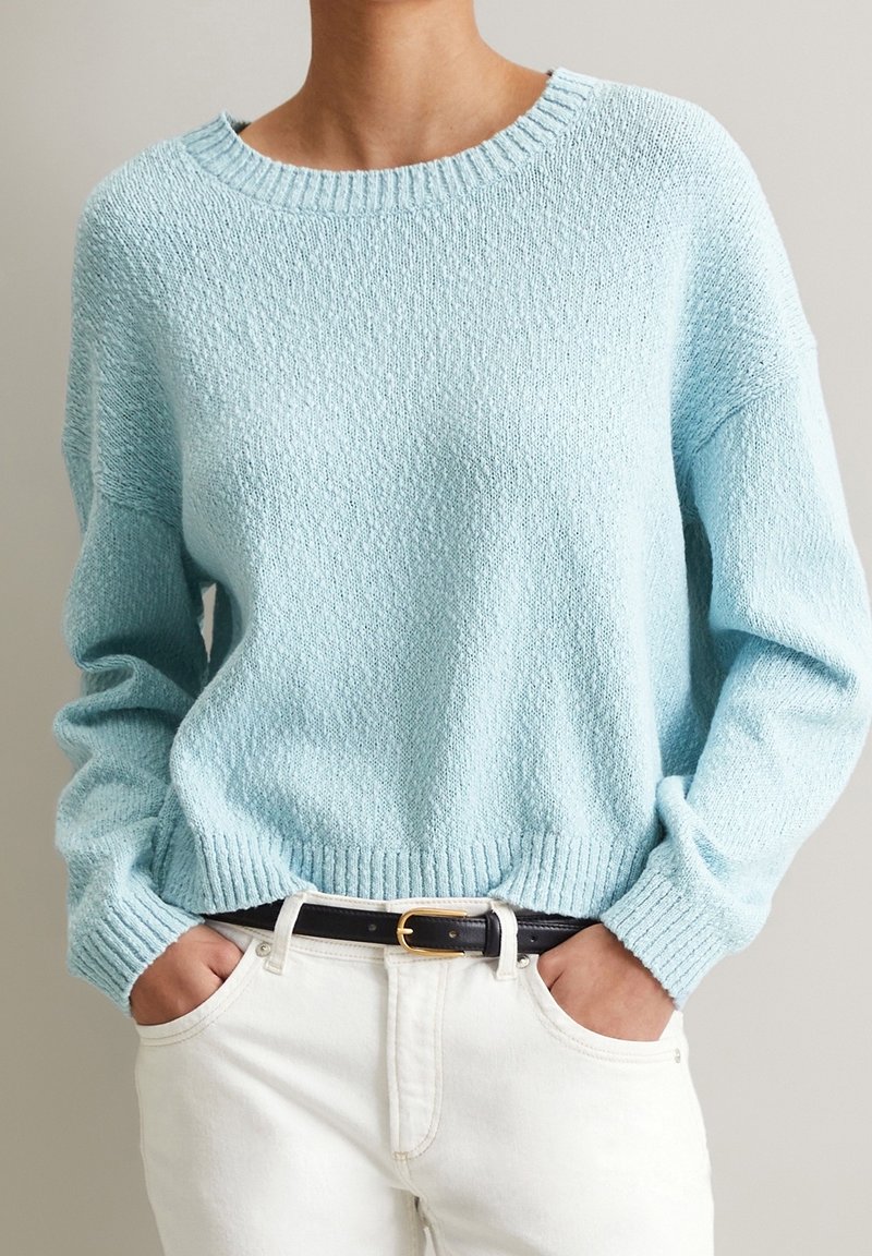 Ljusblå stickad tröja med strukturerad yta, rundad hals och ribbade ärmar. Kombinerad med vita byxor och ett svart bälte.
