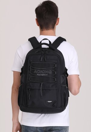 Zaino nero con molteplici scomparti, spallacci regolabili e un logo strutturato sulla parte frontale. Realizzato in tessuto resistente con zip e cordini.