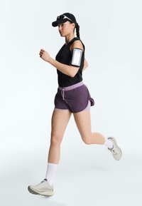 Femme avec une casquette noire, un débardeur et un short violet en train de courir, portant des chaussettes blanches, des chaussures de sport et un brassard avec un support pour téléphone.
