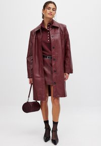 Bordeaux faux leren jas met een glanzende afwerking, grote kraag, zwarte knopen, gecombineerd met een bijpassende rok, hakken en een ronde handtas.