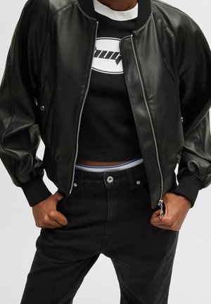 Persona che indossa un bomber in pelle nera sopra una maglietta nera corta con un logo bianco, e jeans neri a vita alta con le mani in tasca.