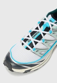 Basket de course blanche avec tige en mesh, accents turquoise, détails réfléchissants et système de laçage noir. Le design comprend une semelle incurvée pour la stabilité.