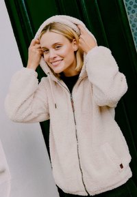 Pehmeast heleda roosa kangast fliisist kapuutsiga jope. Omab tõmbluku sulgemist, küljetaskuid ja tekstuuri. Akrüülideks on pruuni tooni detailid.