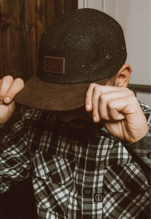 Black cap with speckled pattern, brown suede brim, leather patch logo. Worn with a black and white plaid flannel shirt.