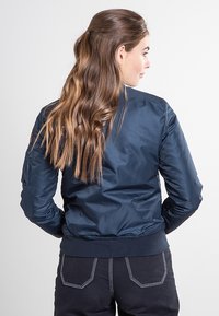 Granatowa kurtka bomberka z gładkiego nylonu, z prążkowanymi mankietami i dołem, ozdobiona naszywką na rękawie, noszona z ciemnymi spodniami.