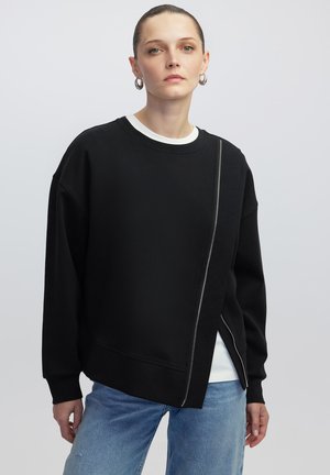 Felpa nera oversize con dettaglio della zip diagonale, dotata di scollo rotondo e una consistenza morbida, abbinata a jeans blu chiaro.