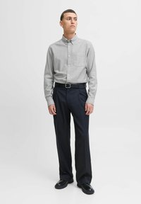 Camicia grigia con bottoni e taschino sul petto, abbinata a pantaloni neri larghi e scarpe nere, caratterizzata da una superficie liscia e un design su misura.