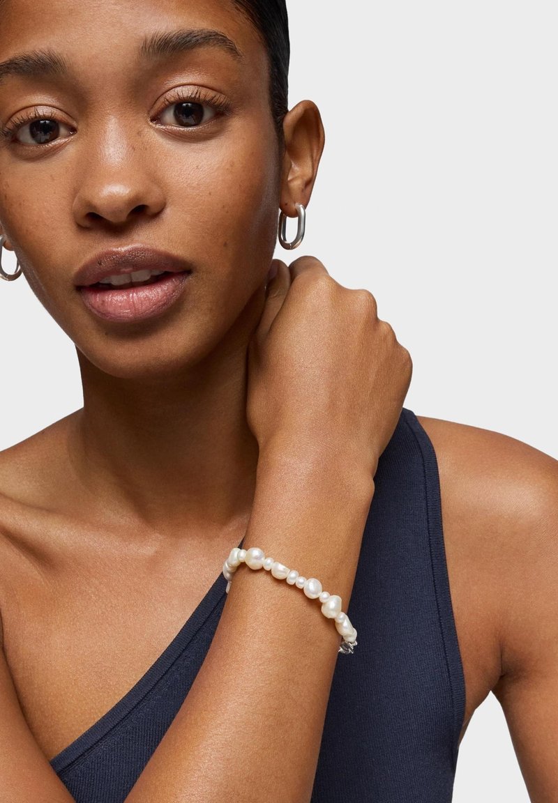 Kobieta nosząca srebrne kolczyki koła, ciemny bezrękawnik i nierówną bransoletkę z białych pereł, trzymająca nadgarstek blisko szyi.