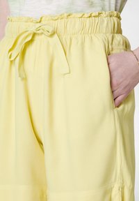 Gula, lätta byxor med elastisk midja och knytning, med sidfickor och en slät textur. Volang i midjan som accent.