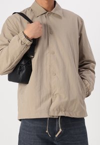 Beżowa lekka kurtka z kołnierzykiem, zatrzaskami i elastycznymi mankietami, zestawiona z małą czarną torebką na ramię.