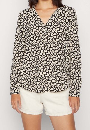 Femme portant un chemisier manches longues noir et blanc à fleurs, associé à un short blanc, visible uniquement du cou au milieu des cuisses.