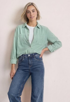 Ung kvinde med kort blondt hår iført en lysegrøn skjorte med knapper over en hvid top og blå jeans med høj talje, der læner sig op ad en væg.