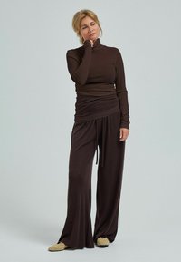 Kvinna i brun polotröja och vida byxor står eftertänksamt med handen på hakan, iförd beige slip-on skor mot en enfärgad bakgrund.