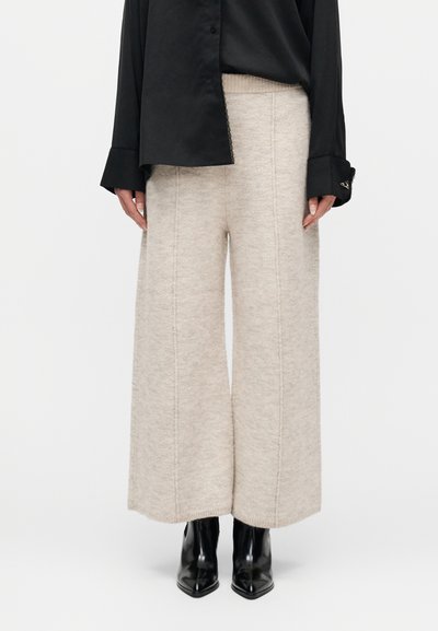 Femme portant un pantalon beige en maille à jambes larges, une chemise noire boutonnée et des bottines noires à bout pointu, debout devant un fond blanc.