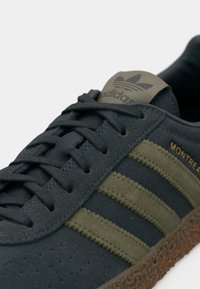 Γκρι σουέντ αθλητικά παπούτσια με ελιά πράσινες ρίγες, διαθέτουν υφή στη σόλα από καουτσούκ και ανάγλυφο λογότυπο στη γλώσσα. Τα κορδόνια είναι μαύρα.