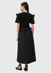 Zwarte jurk met een smock-buste, korte pofmouwen en een gerimpelde zoom. De silhouette is lang en vloeiend, gecombineerd met zwarte sandalen.