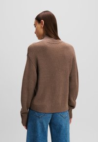 Brauner gerippter Pullover mit hohem Kragen und langen Ärmeln, kombiniert mit lockeren blauen Jeanshosen. Weicher, strukturierter Stoff mit lässiger Passform.