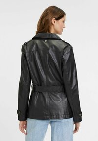 Giacca di pelle nera con vita regolabile, spalline e bottoni sui polsini. Texture liscia e design sartoriale.