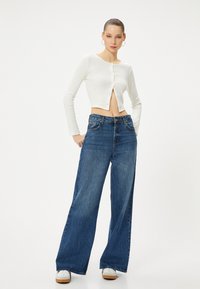 Witte geribbelde lange mouwen crop top met knoopsluiting aan de voorkant, gecombineerd met blauwe wijdlopende denim jeans met een hoge taille en minimale slijtage.