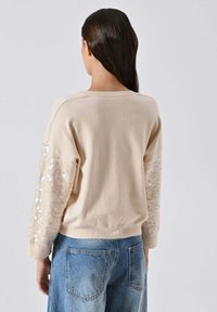 Maglione beige con maniche lavorate a trama e decorate con paillettes. Scollo tondo, orlo a coste e vestibilità casual. Abbinato a jeans in denim azzurro chiaro.