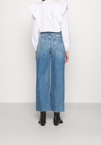 Wide-leg blue denim jeans with a faded finish, featuring a mid-rise waist and classic back pockets, paired with a white blouse with ruffles.