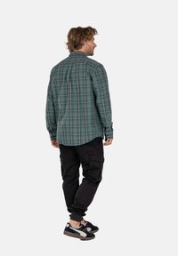 Camisa de cuadros verde de botones y mangas largas, combinada con pantalones cargo negros y zapatillas deportivas. Detalles de costura visibles y dobladillo redondeado.