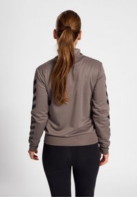 Brown athletic jacket with a high collar, featuring black striped accents on sleeves and a snug waistband. Smooth texture, fitted design.