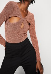 Brun glittrig långärmad topp med utskärningar och knutdetaljer, bärs tillsammans med svarta höga byxor och håller i en svart kuvertväska.