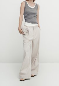Grå tætsiddende tanktop lagdelt over en hvid tanktop, parret med løse beige bukser. Teksturen er glat, med flad front og vide ben. Holder en beige clutch.