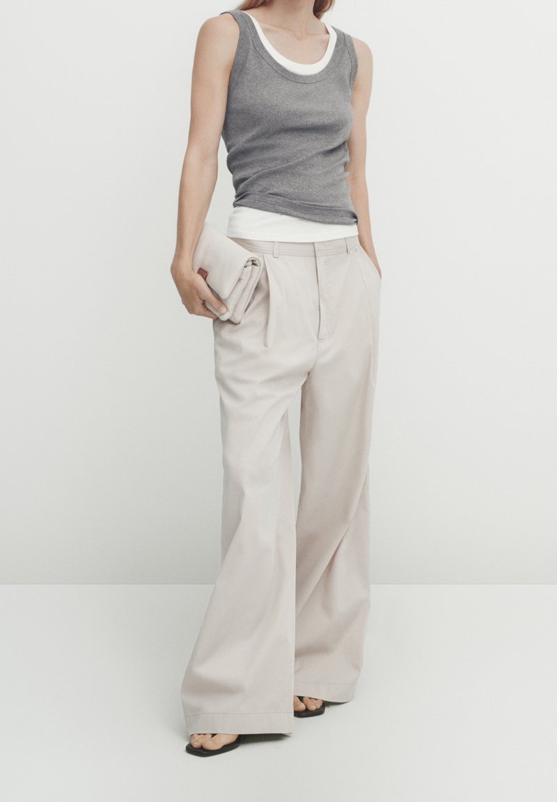 Grå tætsiddende tanktop lagdelt over en hvid tanktop, parret med løse beige bukser. Teksturen er glat, med flad front og vide ben. Holder en beige clutch.