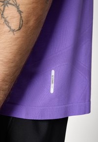 Lila Sportshirt mit Mesh-Textur, geripptem Saum und "ACTIBREEZE"-Etikett an der unteren Seite. Getragen mit schwarzen Hosen.