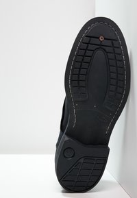 Semelle de chaussure en caoutchouc noir avec un motif de grip texturé, un détail de disque circulaire et des coutures blanches. Vue en angle sur fond clair.