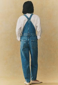 Denim tuinbroek in lichtblauw, met verstelbare banden, een comfortabele pasvorm en achterzakken, gecombineerd met een eenvoudig wit shirt en beige flats.
