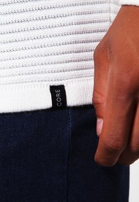 Maglione bianco a coste con un motivo a maglia testurizzata, dotato di un'etichetta nera cucita all'orlo. Abbinato a jeans blu scuro.