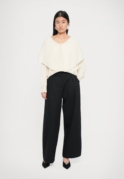 Pull en maille crème avec un col large drapé, associé à un pantalon noir à jambes larges. Le mannequin porte des escarpins noirs à bouts pointus et de grandes boucles d'oreilles.