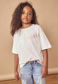 Šviesiai rožinė marškinėliai su užsiūtu „SAKURA“ tamsesnės rožinės spalvos, derinami su išblukusiomis mėlynomis suplėšytomis džinsais. Minkšta medžiaga, laisvas kirpimas, trumpi rankoviai.