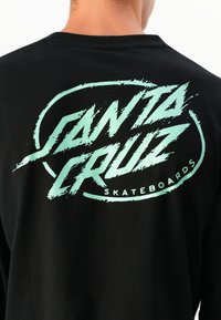 Zwarte langwerpige shirt met lange mouwen, featuring een groot mintgroen grafisch tekstontwerp op de achterkant met de tekst "SANTA CRUZ SKATEBOARDS".