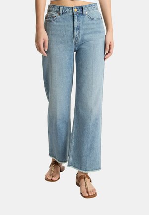 Persona che indossa jeans azzurri a gamba larga con orli grezzi e sandali infradito marroni con dettagli in catena dorata.